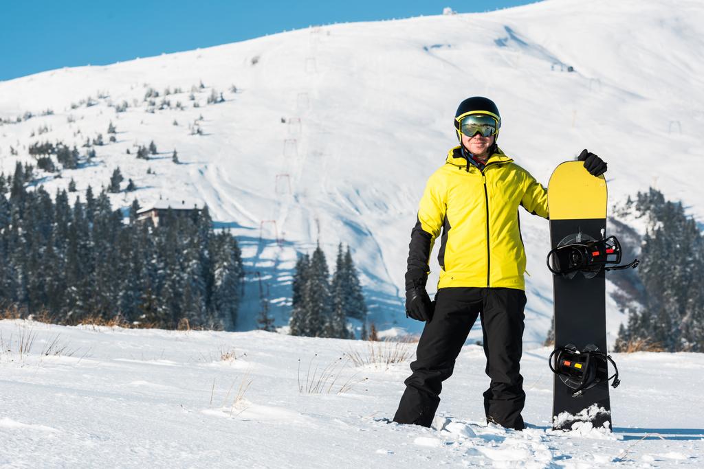 fröhlicher Sportler mit Helm und Snowboard auf weißem Schnee in den Bergen - Foto, Bild
