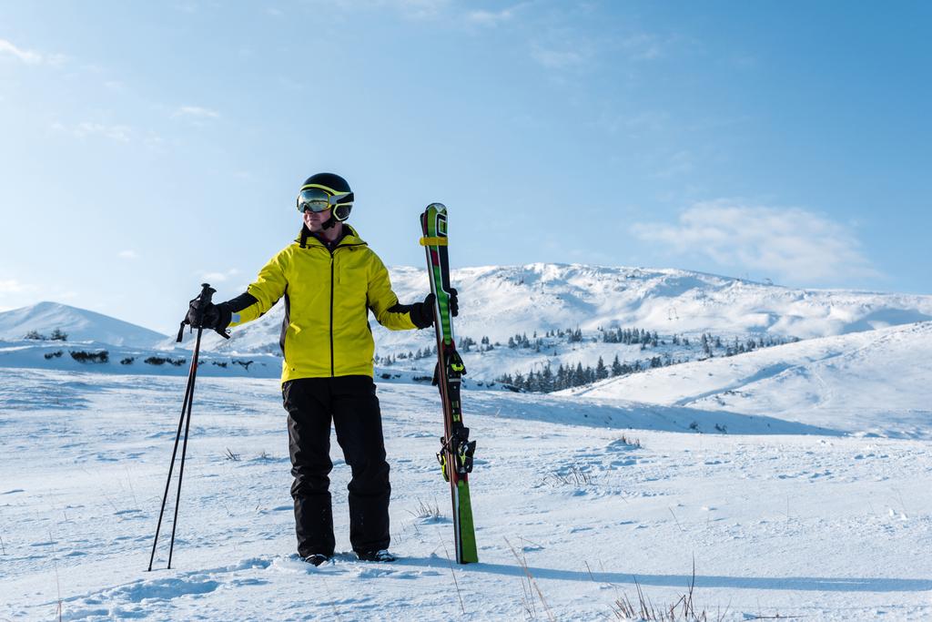 σκιέρ σε κράνος και γυαλιά στέκεται με μπαστούνια σε λευκό χιόνι στα βουνά - Φωτογραφία, εικόνα