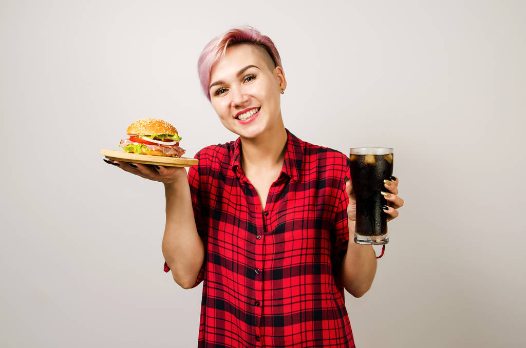 Joven hermosa chica sostiene hamburguesa y vaso de cola, sobre un fondo blanco
. - Foto, Imagen