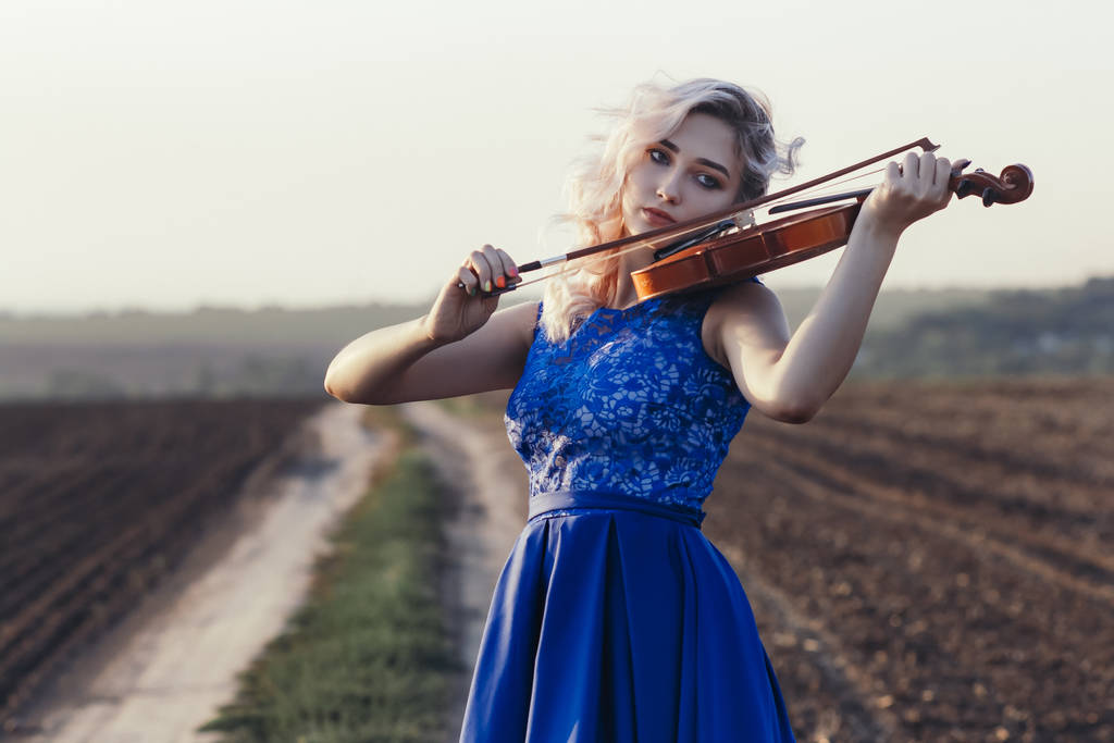 kobieta w długiej sukience grająca na skrzypcach na tle polnej ścieżki z klubem kurzu, dziewczyna zajmująca się sztuką muzyczną, performans na łonie natury, koncepcja pasji w muzyce - Zdjęcie, obraz