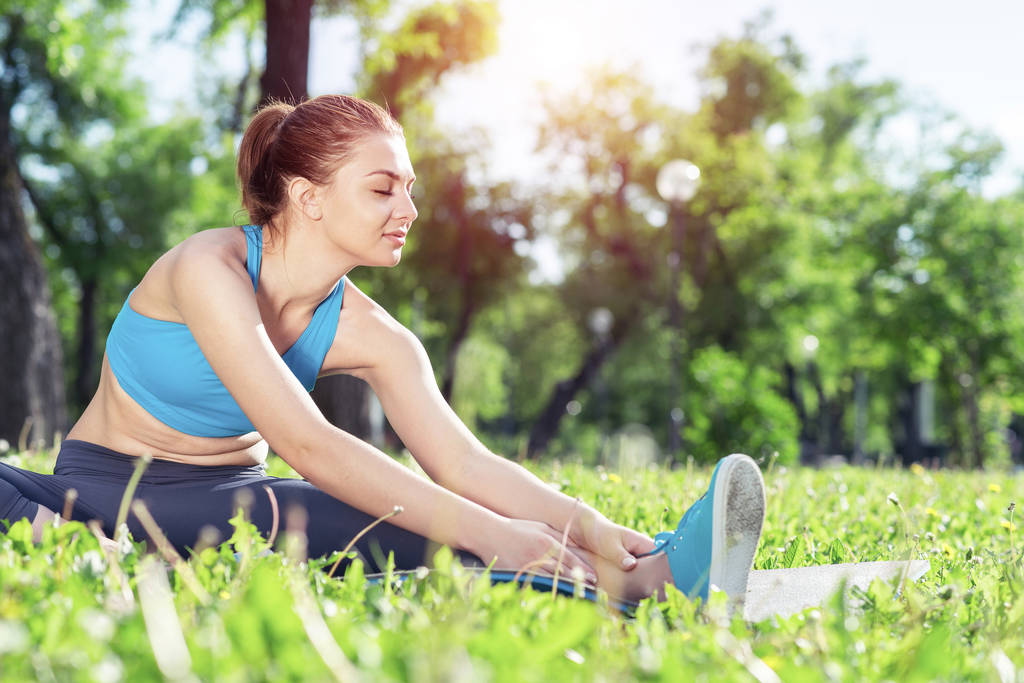 Krásná usměvavá dívka protáhnout před fitness trénink v parku při východu slunce. Mladá žena v kombinéze sedí v letní sezóně na zelené trávě. Ranní cvičení a zdravý životní styl. - Fotografie, Obrázek