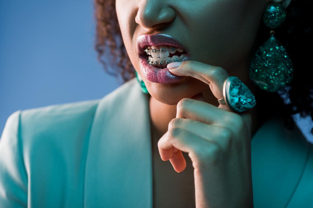 przycięty widok afrykańskiej amerykanki z aparatem na zębach gryzący palec odizolowany na niebiesko - Zdjęcie, obraz
