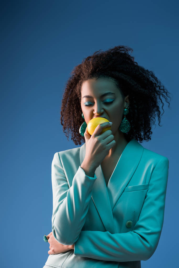 mujer afroamericana con estilo con maquillaje comer limón aislado en azul
 - Foto, Imagen