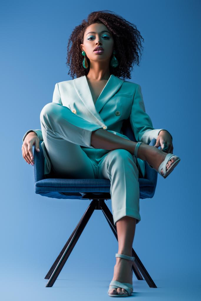 Afro-Amerikan kadın mavi arka planda koltukta oturuyor.  - Fotoğraf, Görsel