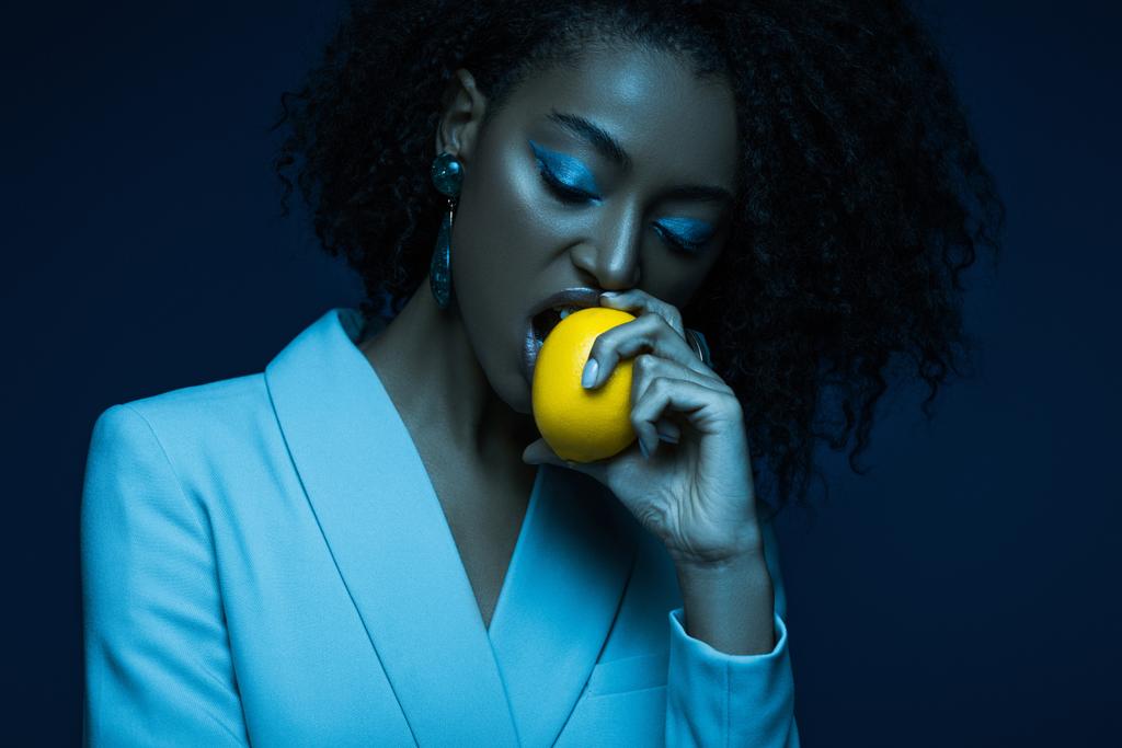 atractiva mujer afroamericana con maquillaje comer limón aislado en azul
 - Foto, Imagen