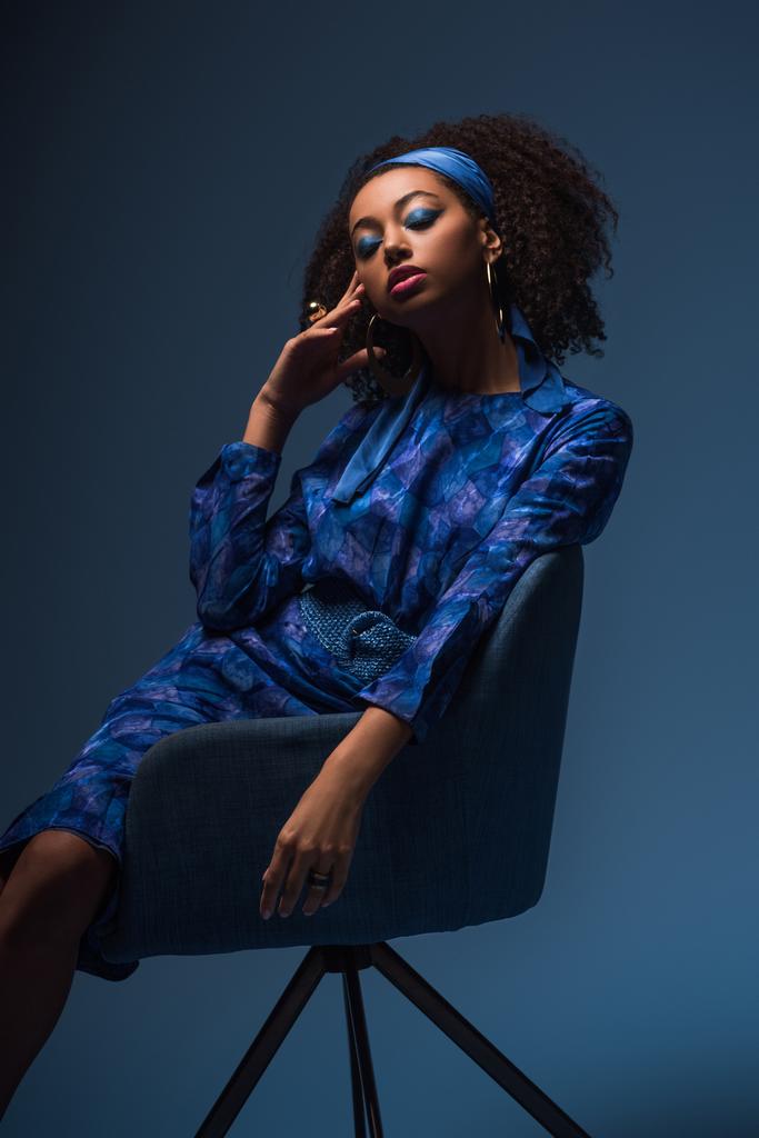 ελκυστική Αφροαμερικανή γυναίκα με κλειστά μάτια κάθεται σε πολυθρόνα απομονωμένη στο μπλε  - Φωτογραφία, εικόνα