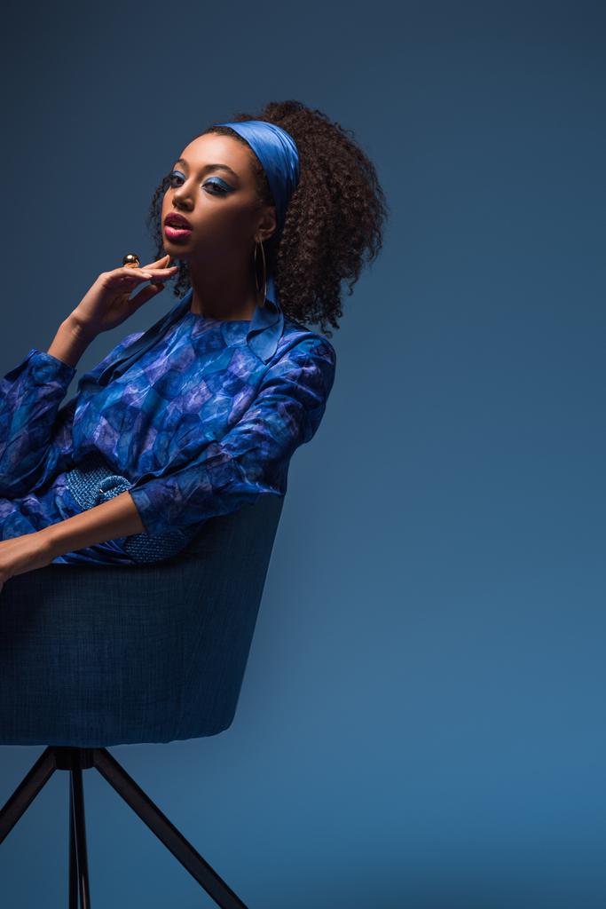 attraktive afrikanisch-amerikanische Frau sitzt auf Sessel isoliert auf blau  - Foto, Bild