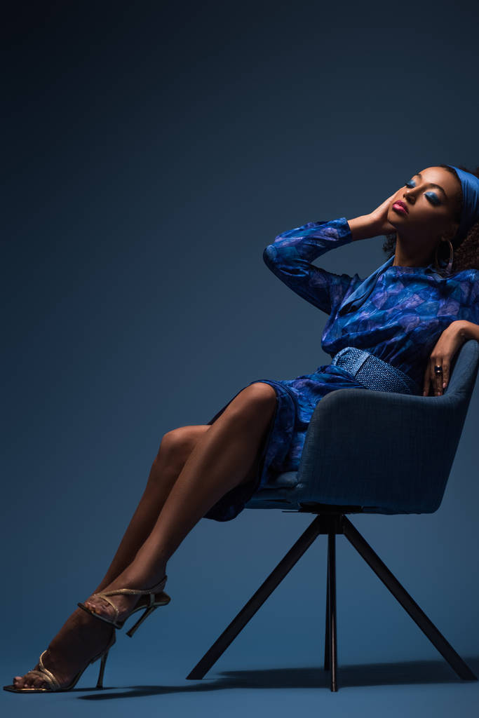 青い背景にアームチェアに座って目を閉じた魅力的なアフリカ系アメリカ人女性  - 写真・画像