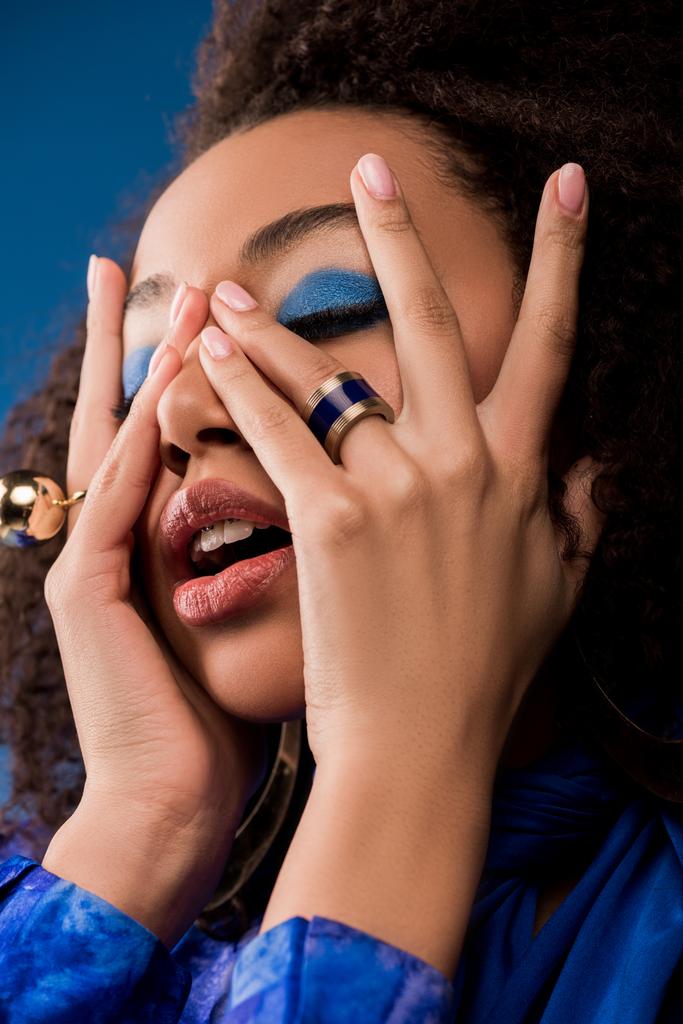 stylowa Afroamerykanka z zamkniętymi oczami zasłaniająca twarz odizolowana na niebiesko   - Zdjęcie, obraz