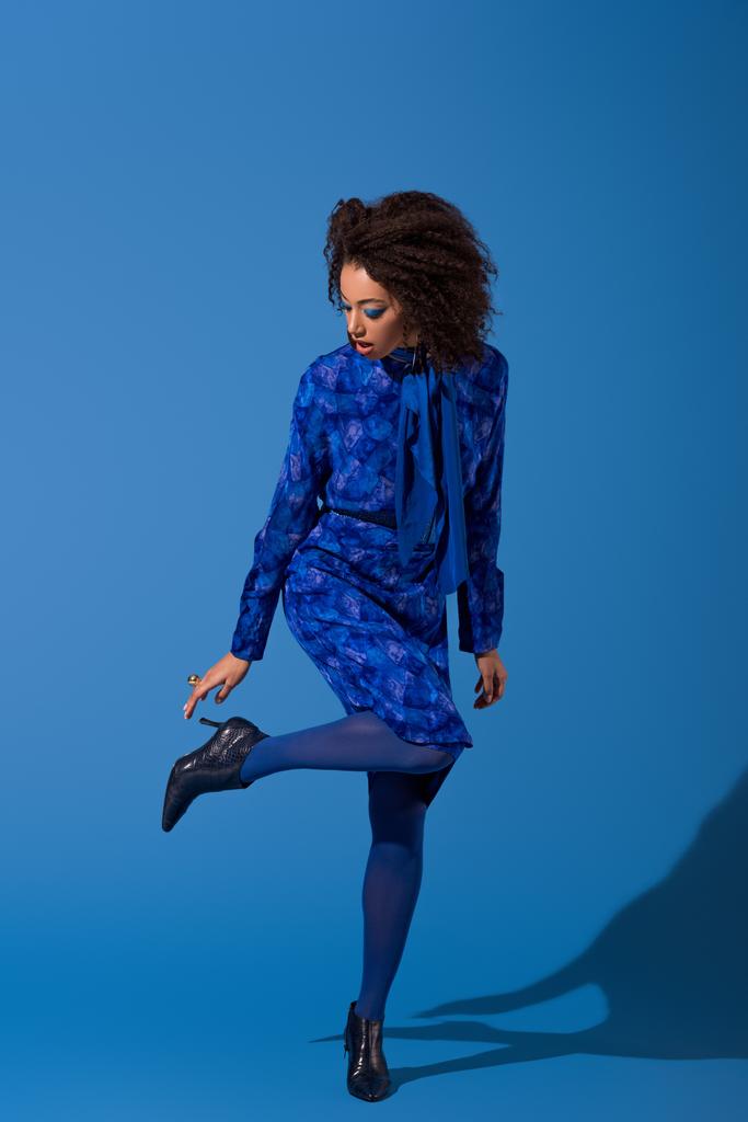 attractive african american woman in dress touching her footwear on blue background  - Photo, Image