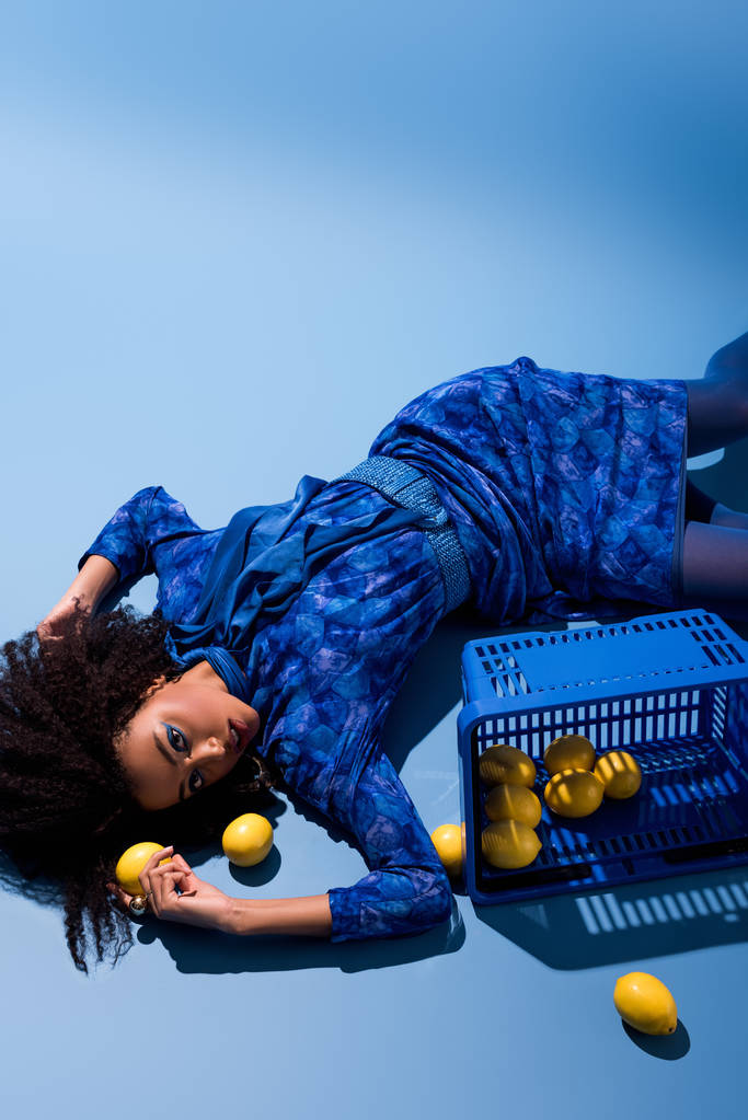 african american woman lying with shopping basket and lemons on blue background  - Фото, зображення