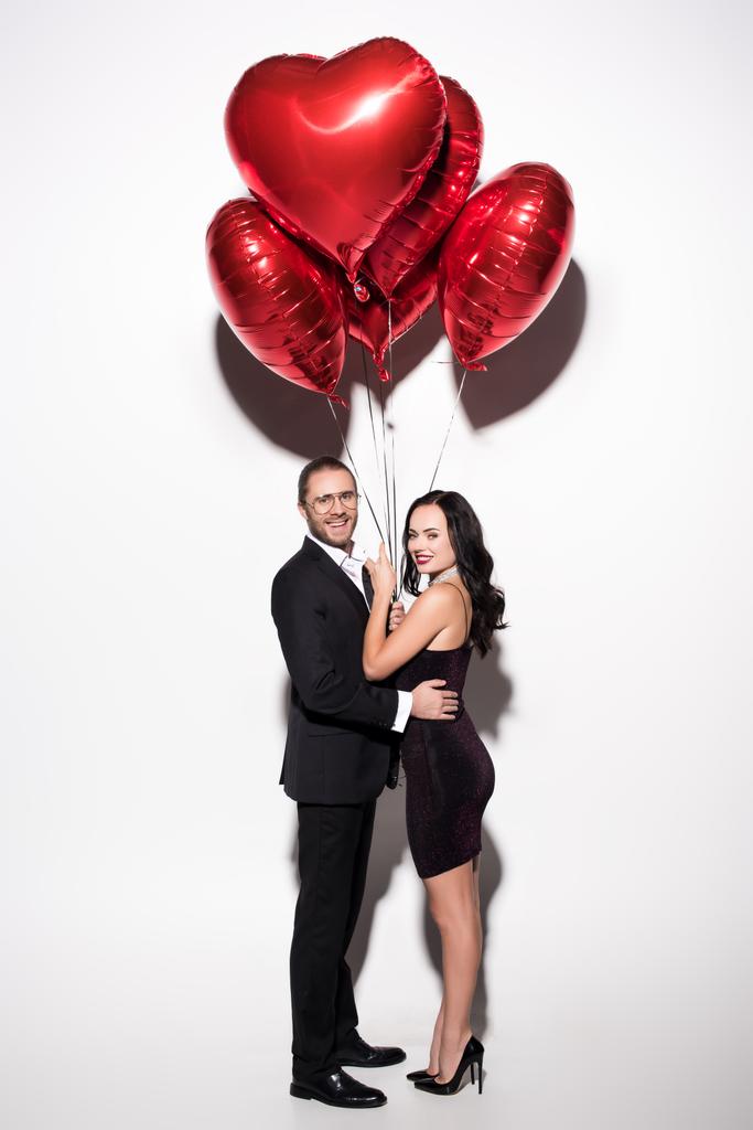 χαμογελαστό ζευγάρι κρατώντας κόκκινα μπαλόνια σχήμα καρδιάς για την ημέρα του Αγίου Βαλεντίνου σε λευκό - Φωτογραφία, εικόνα