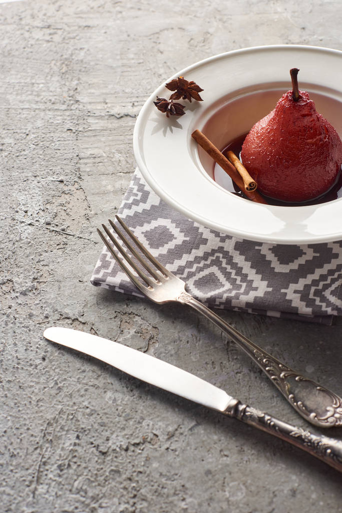 вкусная груша в красном вине с корицей и анисом на тарелке на серой бетонной поверхности с столовыми приборами на салфетке
 - Фото, изображение