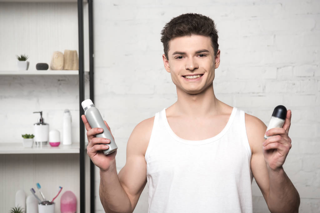 Beyaz kolsuz gömlekli genç adam deodorant tutuyor ve kameraya gülümsüyor. - Fotoğraf, Görsel