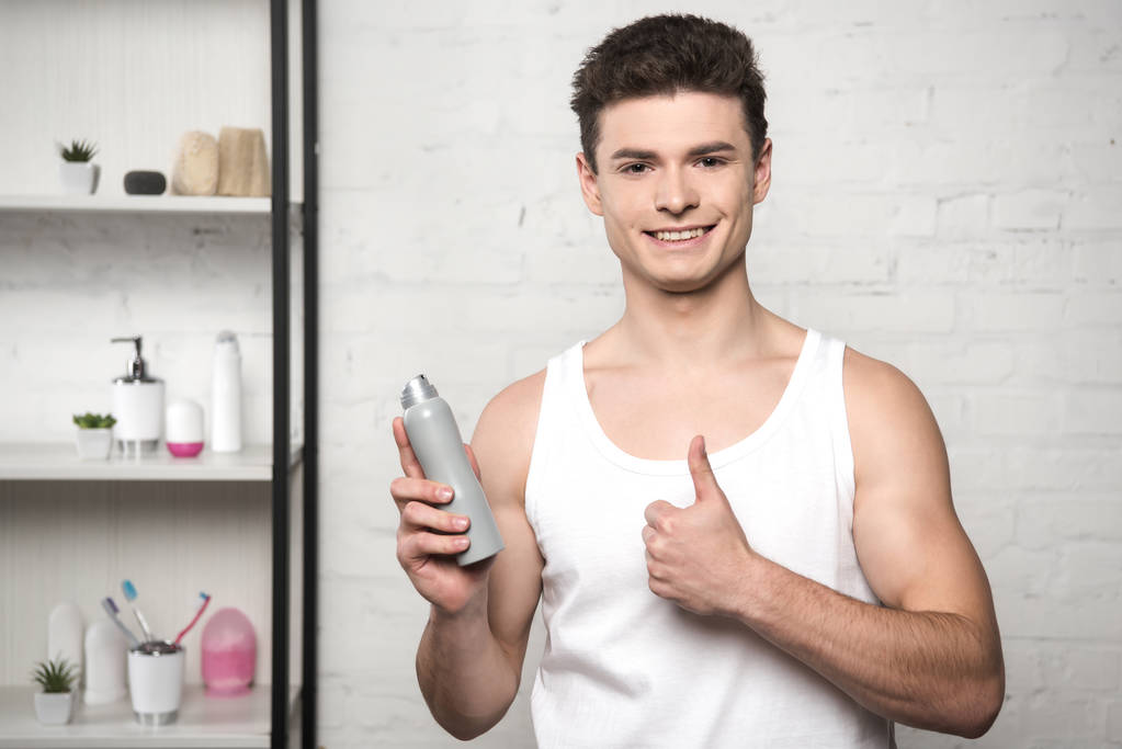 usmívající se muž v bílé košili bez rukávů ukazující palec nahoru, zatímco drží deodorant - Fotografie, Obrázek