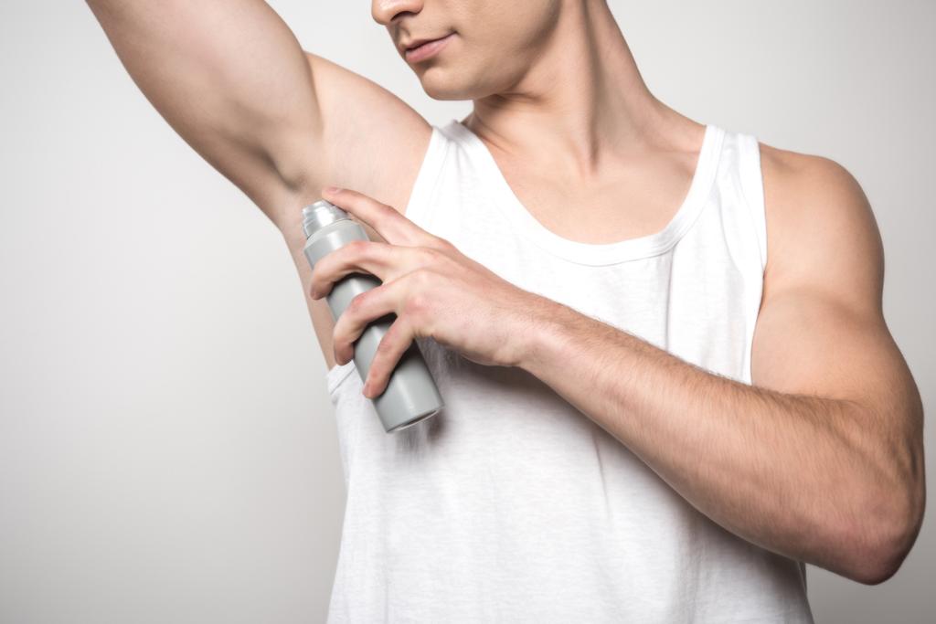 Beyaz kolsuz gömlekli genç bir adamın kolunun altına deodorant sürerken görüntüsü gri üzerine izole edilmiş. - Fotoğraf, Görsel