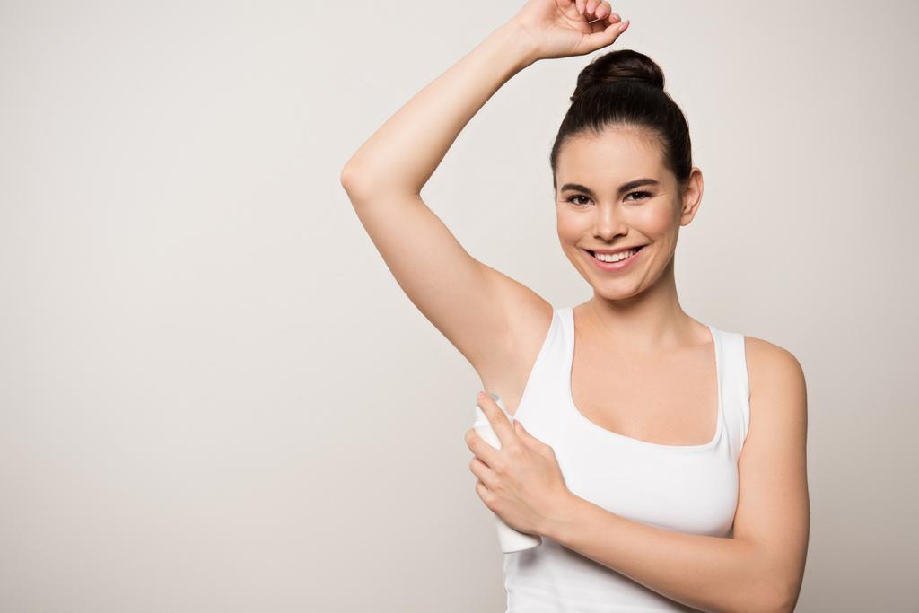 vrolijke vrouw glimlachen op camera terwijl het aanbrengen van deodorant op de onderarm geïsoleerd op grijs - Foto, afbeelding