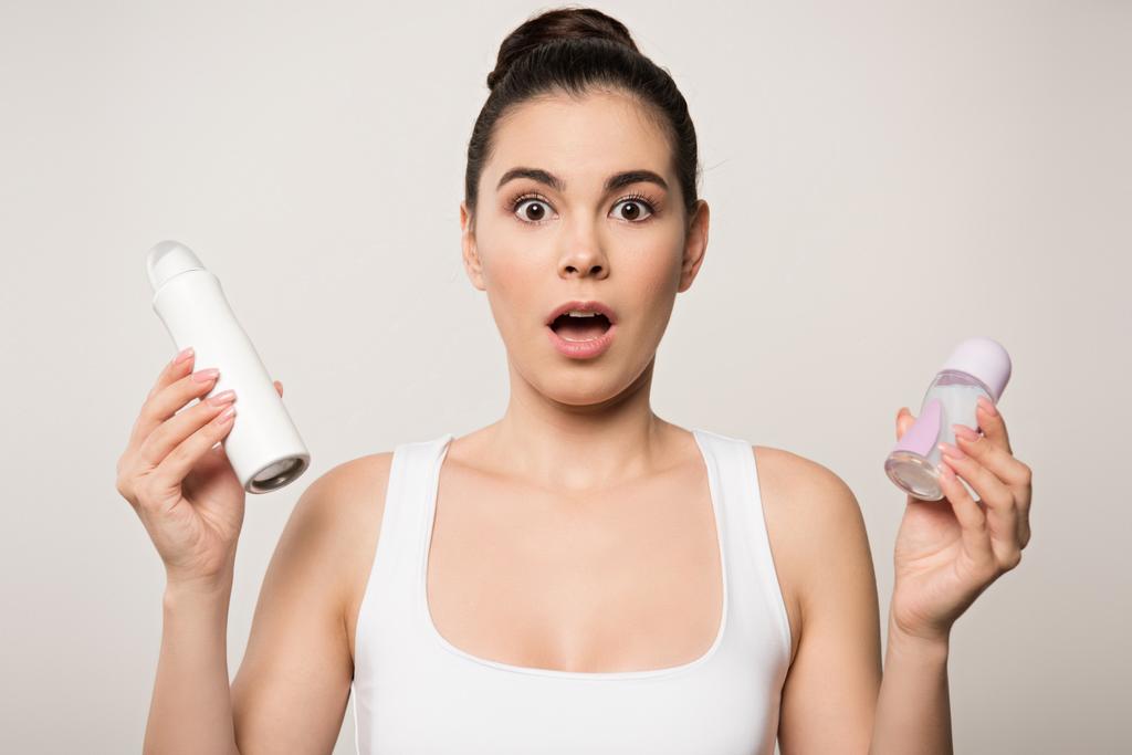 взволнованная женщина смотрит в камеру, держа в руках дезодоранты, изолированные на сером
 - Фото, изображение