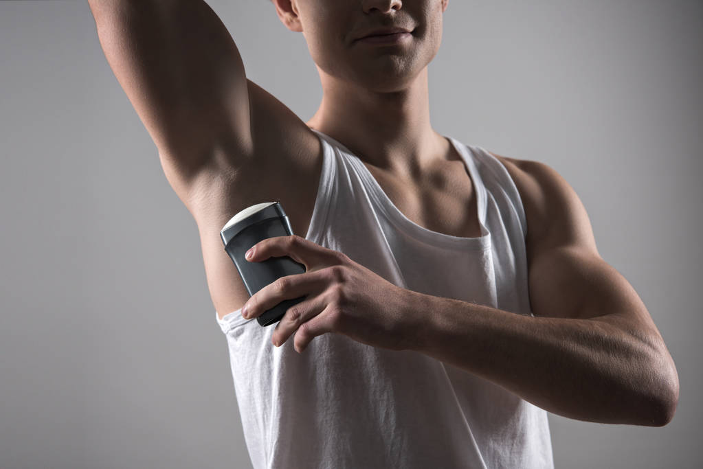 rajattu näkymä nuori mies valkoinen hihaton paita soveltamalla deodorantti kainalossa eristetty harmaa
 - Valokuva, kuva