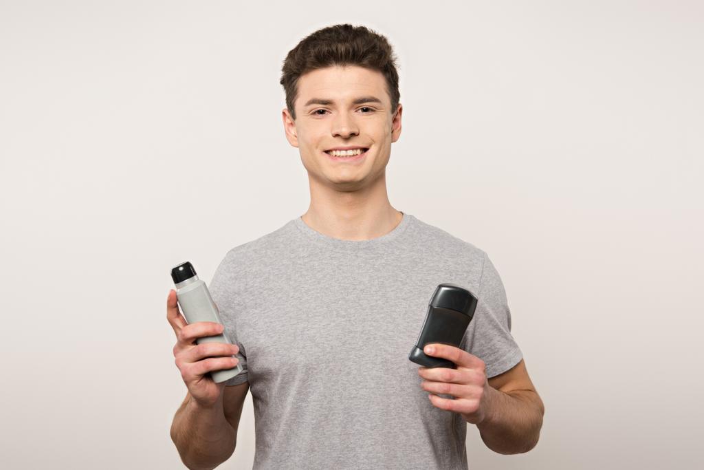 молодий чоловік у сірій футболці тримає дезодоранти і посміхається на камеру ізольовано на сірому
 - Фото, зображення