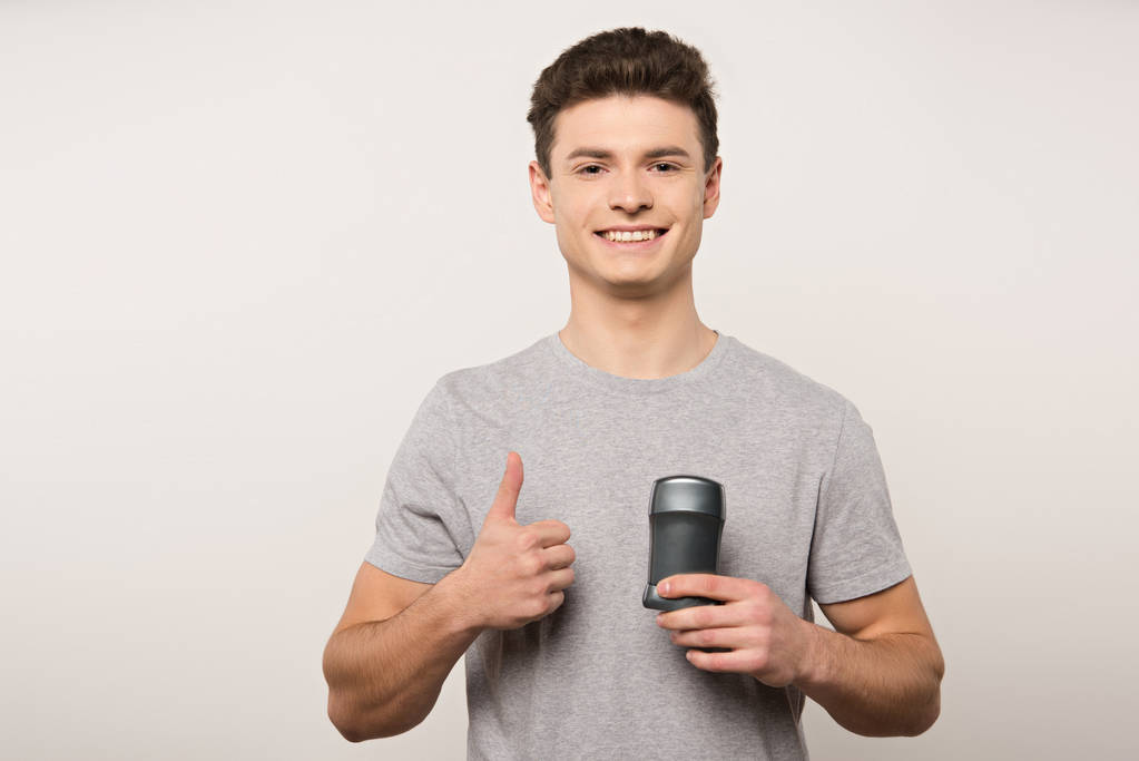 hombre sonriente en camiseta gris sosteniendo desodorante y mostrando el pulgar hacia arriba aislado en gris
 - Foto, Imagen