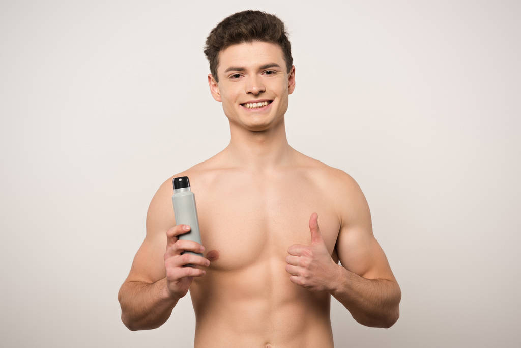 usmívající se muž bez košile ukazující palec nahoru, zatímco drží deodorant izolovaný na šedi - Fotografie, Obrázek