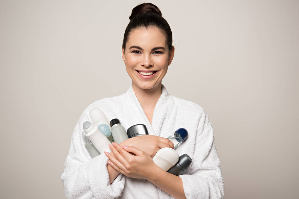 iloinen nainen kylpytakissa, jolla on erilaisia deodorantteja katsoessaan kameraa, joka on eristetty harmaalla
 - Valokuva, kuva