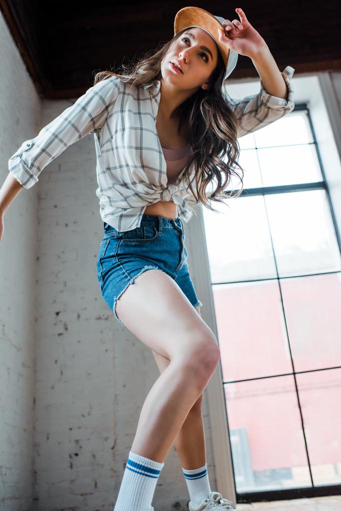 άποψη χαμηλής γωνίας του ελκυστική χορεύτρια χορό hip-hop και αγγίζοντας το καπέλο στο στούντιο χορού  - Φωτογραφία, εικόνα