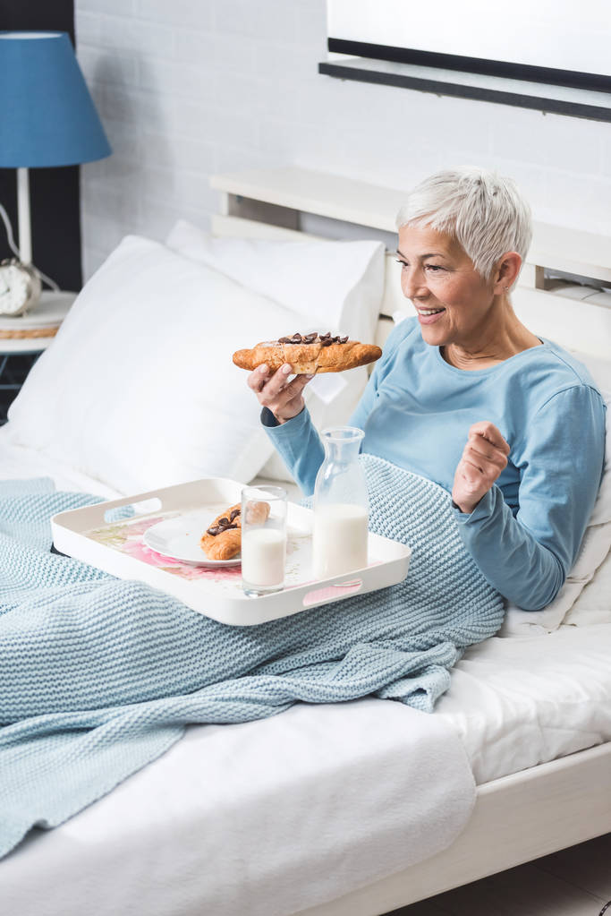 Vanhempi nainen syö aamiaista sängyssä katsomassa televisiota ja syömässä suklaata croissant
 - Valokuva, kuva