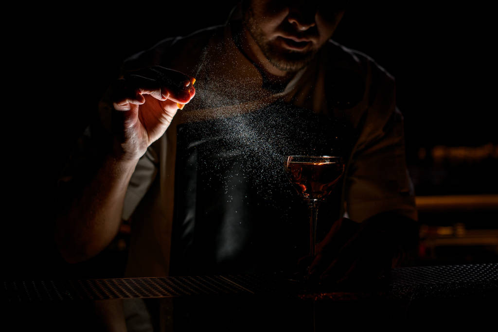 Επαγγελματίας μπάρμαν ψεκάζει το καφέ αλκοολούχο ποτό με χυμό ξύσμα πορτοκαλιού στο σκοτάδι - Φωτογραφία, εικόνα