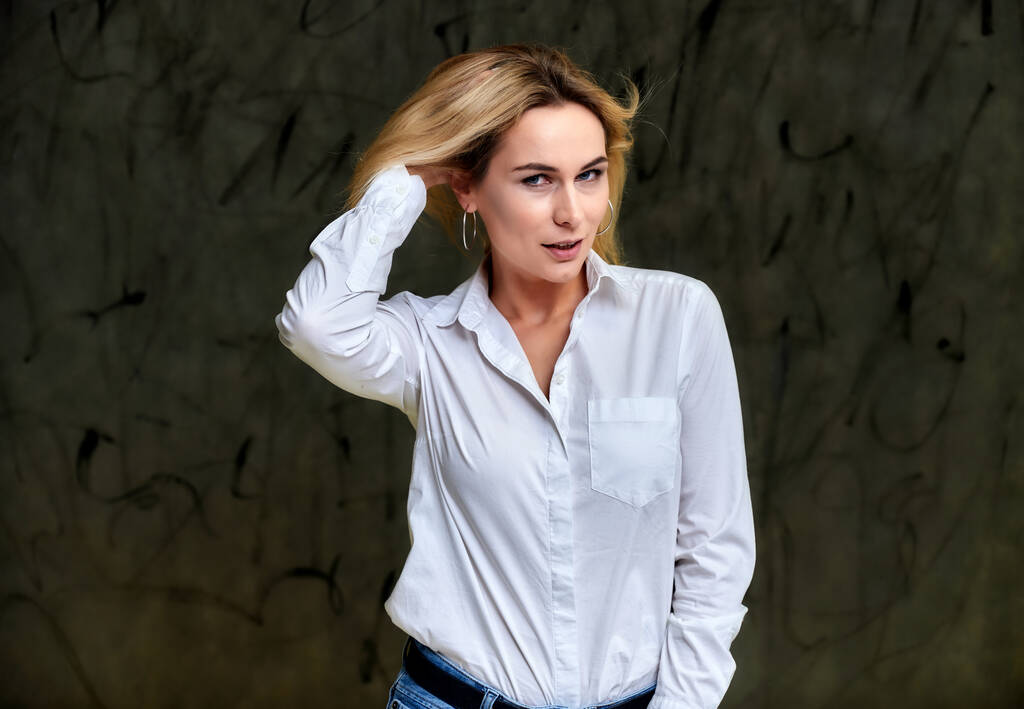 Retrato de uma mulher loira muito sorridente em uma camisa branca com cabelo voador em pé sobre um fundo alternativo cinza. Bem na frente da câmera com emoções
. - Foto, Imagem