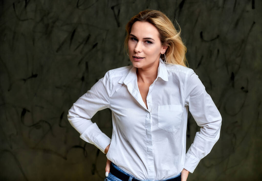 Retrato de uma mulher loira muito sorridente em uma camisa branca com cabelo voador em pé sobre um fundo alternativo cinza. Bem na frente da câmera com emoções
. - Foto, Imagem