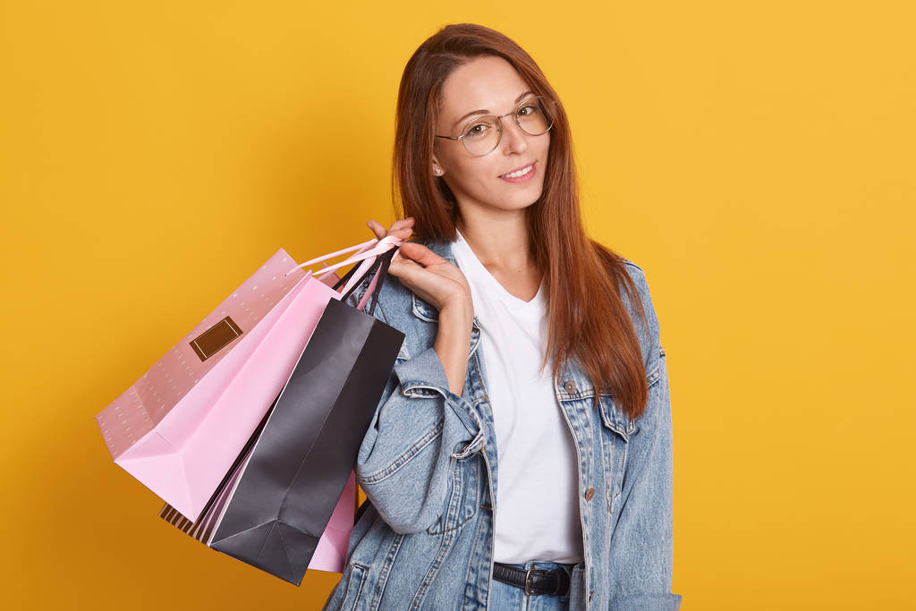 Porträt einer schönen Frau mit glatten Haaren, die rosa und schwarze Einkaufstüten vor gelbem Studiohintergrund hält, direkt in die Kamera blickt, lässige Kleidung und Brille trägt. - Foto, Bild