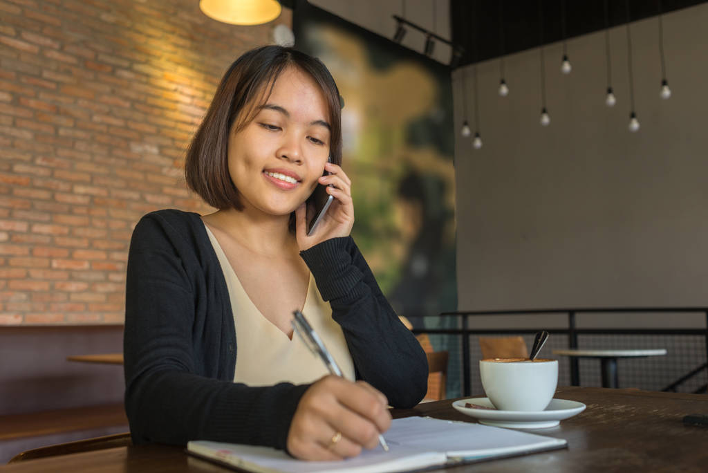 Aziatische dame schrijft notities, beantwoordt telefoongesprek in de coffeeshop - Foto, afbeelding