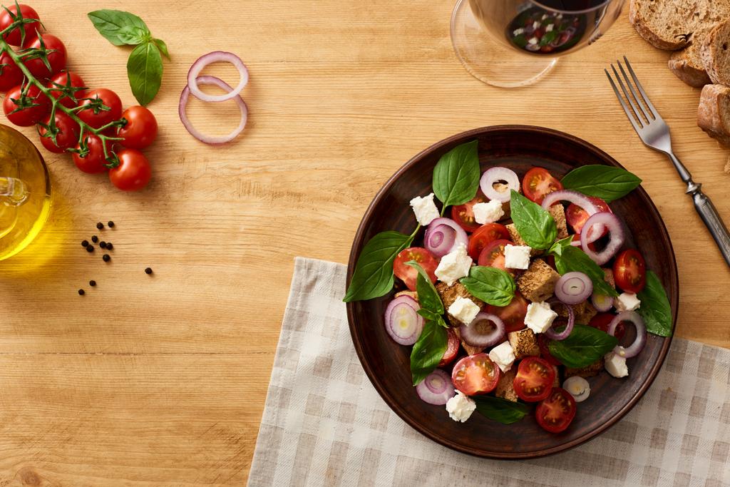 vista superior de la deliciosa ensalada de verduras italiana panzanella servido en el plato en la mesa de madera cerca de ingredientes frescos y vino tinto
 - Foto, Imagen