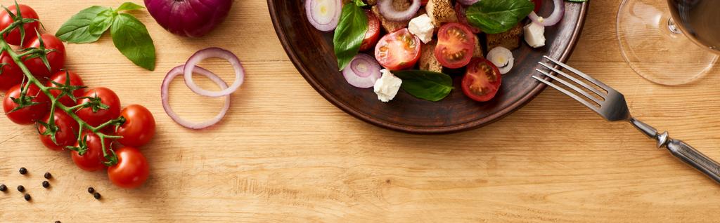 сверху вид на вкусный итальянский овощной салат panzanella подается на тарелке с вилкой на деревянном столе рядом со свежими ингредиентами и красным вином, панорамный снимок
 - Фото, изображение