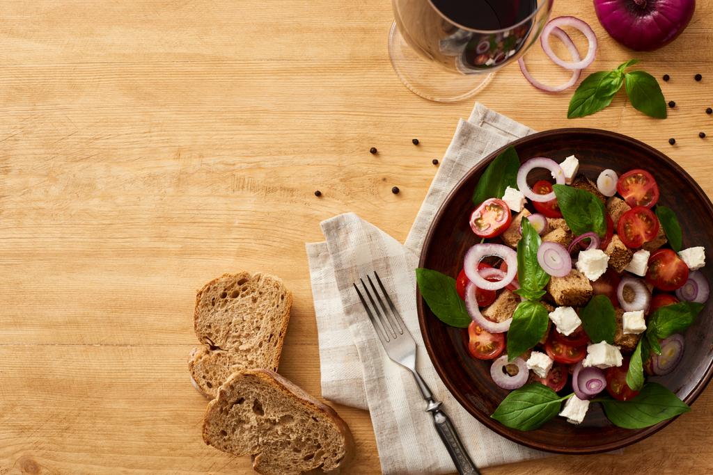 τοπ άποψη των νόστιμα ιταλική σαλάτα λαχανικών panzanella σερβίρεται στο πιάτο σε ξύλινο τραπέζι κοντά φρέσκα υλικά, πιρούνι, ψωμί και κόκκινο κρασί - Φωτογραφία, εικόνα