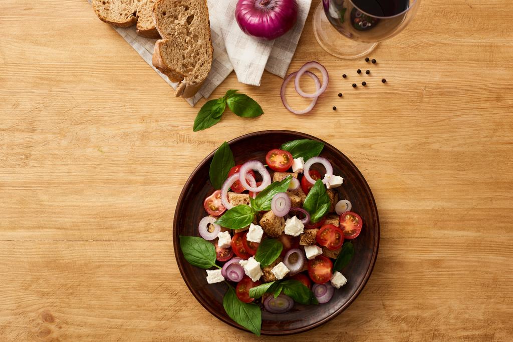 τοπ άποψη των νόστιμα ιταλική σαλάτα λαχανικών panzanella σερβίρεται στο πιάτο σε ξύλινο τραπέζι κοντά φρέσκα υλικά σε χαρτοπετσέτα και κόκκινο κρασί - Φωτογραφία, εικόνα
