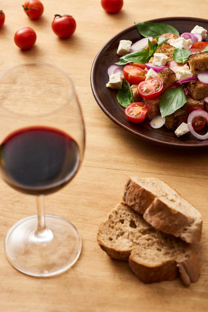 selektiver Schwerpunkt von köstlichem italienischem Gemüsesalat Panzanella, serviert auf Teller auf Holztisch in der Nähe von Rotwein und Brot - Foto, Bild