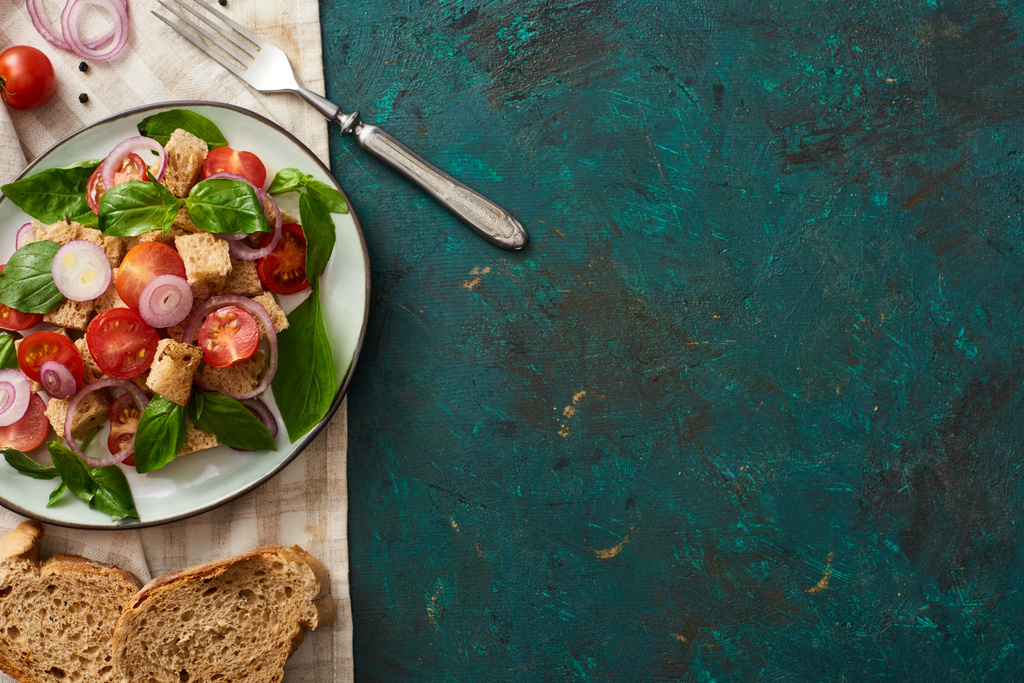 vrchní pohled na vynikající italský zeleninový salát panzanella podávané na talíři na texturovaném zeleném povrchu s chlebem, ingrediencemi, ubrouskem a vidličkou - Fotografie, Obrázek