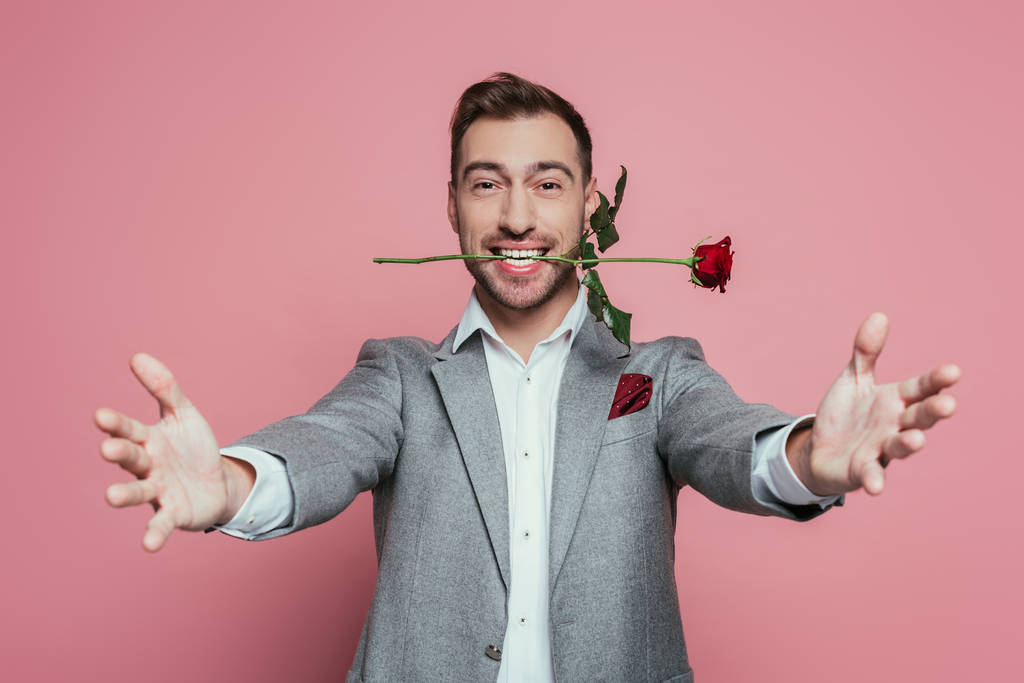 šťastný muž v obleku držící rudou růži v zubech s otevřenou náručí, izolovaný na růžové - Fotografie, Obrázek