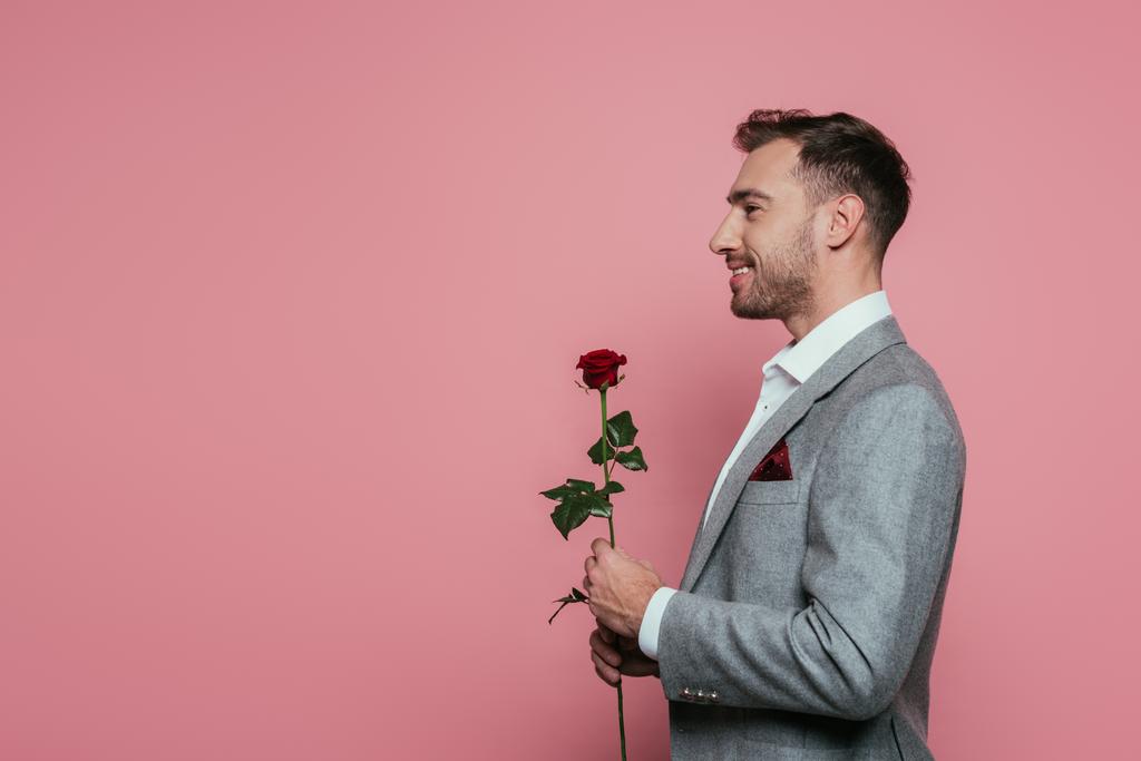 χαμογελαστός γενειοφόρος άνδρας με κοστούμι κρατώντας κόκκινο τριαντάφυλλο, απομονωμένος σε ροζ - Φωτογραφία, εικόνα