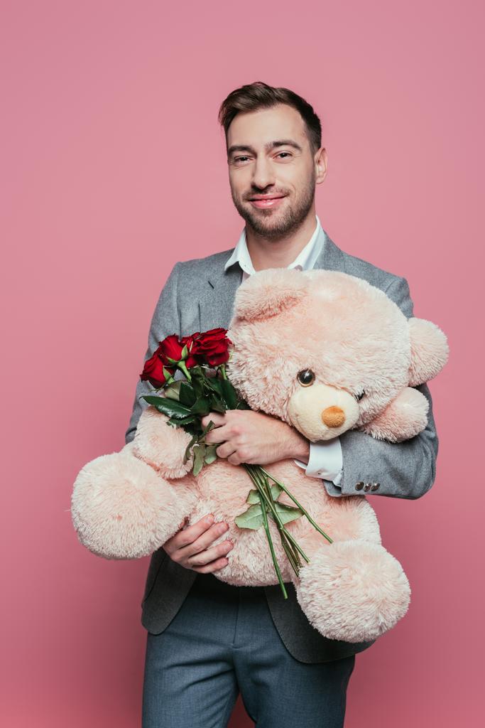 όμορφος χαρούμενος άντρας που κρατάει αρκουδάκι και κόκκινα τριαντάφυλλα, απομονωμένος στο ροζ - Φωτογραφία, εικόνα