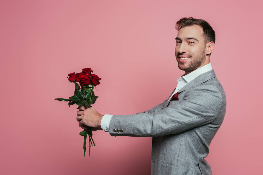 Красивый веселый мужчина в костюме держит красные розы, изолированные на розовый
 - Фото, изображение