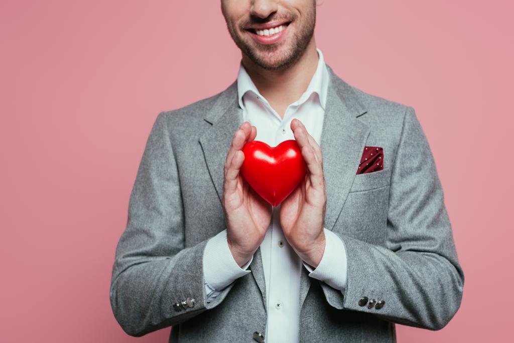 przycięty widok uśmiechniętego mężczyzny trzymającego czerwone serce na Walentynki, odizolowany na różowo  - Zdjęcie, obraz