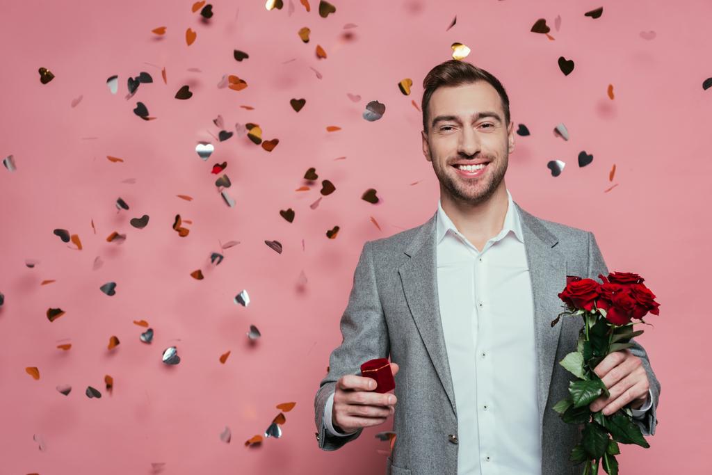 sonriente hombre sosteniendo rosas y anillo de propuesta en rosa con corazones de confeti
 - Foto, imagen