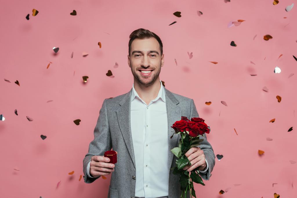 cheerful man holding proposal ring and rose flowers on pink with confetti hearts - Photo, Image