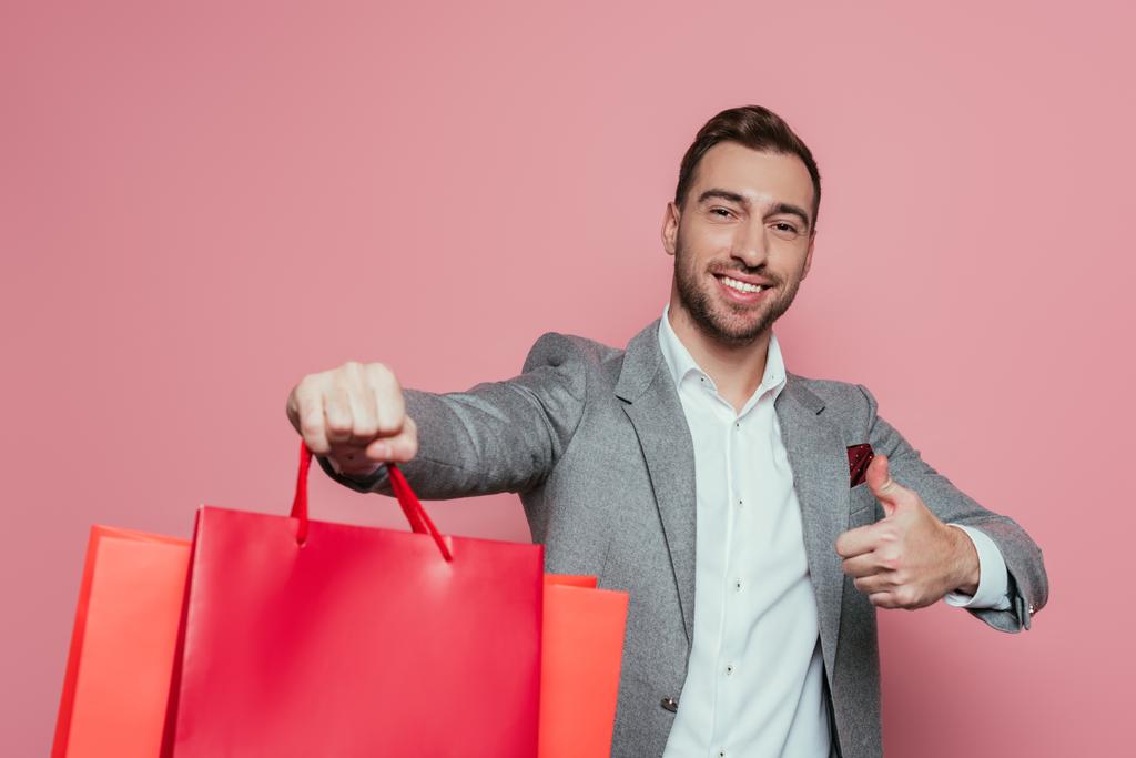 uśmiechnięty mężczyzna trzymający torby na zakupy i pokazujący kciuk w górze, odizolowany na różowo - Zdjęcie, obraz