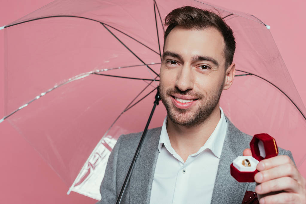χαρούμενο άνδρα κρατώντας δαχτυλίδι πρόταση και ομπρέλα, απομονωμένο σε ροζ  - Φωτογραφία, εικόνα