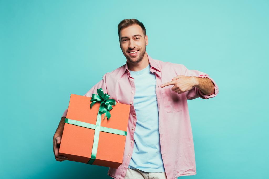 Gülümseyen adam mavi üzerine izole edilmiş büyük hediye kutusunu işaret ediyor. - Fotoğraf, Görsel